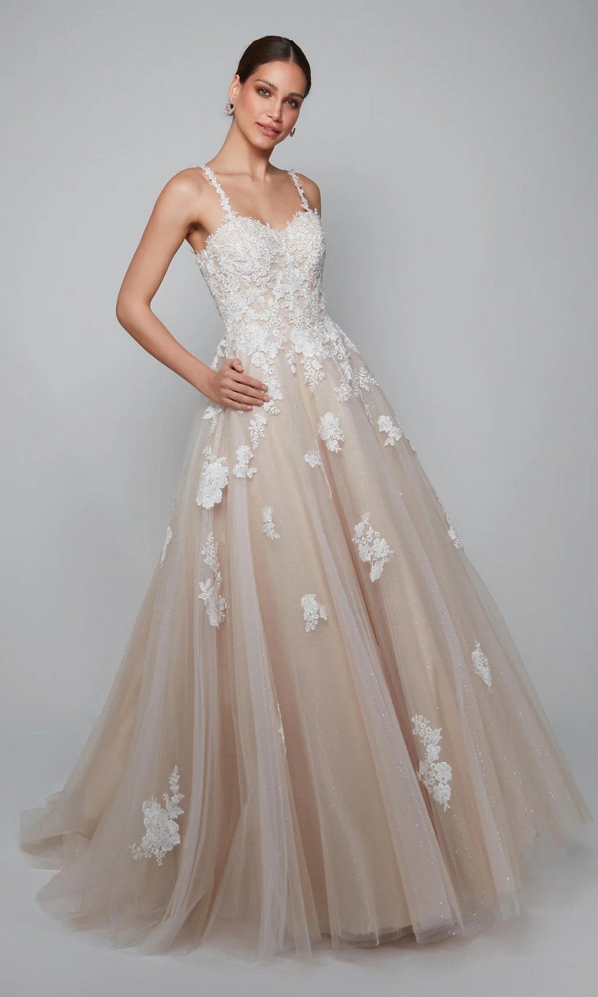 A woman wears the Alyce 7084 Long White Dress featuring white floral lace appliqués, thin straps, a fitted bodice, and a graceful silhouette, standing against a plain gray background.